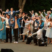Freilichtbühne in Bökendorf Aufführung auf einer Freilichtbühne mit einer großen Gruppe in farbenfrohen Kostümen und erhobenen Armen.