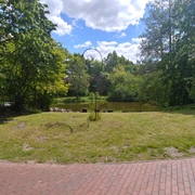 Füllhorn oder Figura Skulptur in einem grünen Park vor einem Teich, umgeben von Bäumen und blauem Himmel mit Wolken.