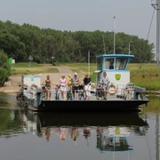 Fähre Schweringen Fähre überquert einen ruhigen Fluss, beladen mit Radfahrern, umgeben von grüner Landschaft.