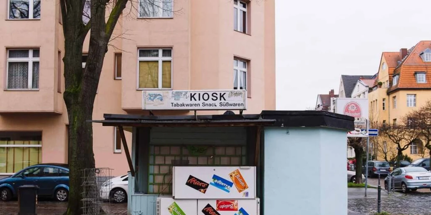Kiosk at Nikolausplatz Kleiner Kiosk auf einer ruhigen Straßenecke, umgeben von Wohngebäuden, mit Werbetafeln für Snacks.Small kiosk on a quiet street corner surrounded by residential buildings, featuring advertising boards for snacks.
