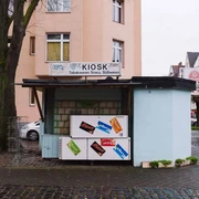 Kiosk at Nikolausplatz Kleiner Kiosk auf einer ruhigen Straßenecke, umgeben von Wohngebäuden, mit Werbetafeln für Snacks.Small kiosk on a quiet street corner surrounded by residential buildings, featuring advertising boards for snacks.