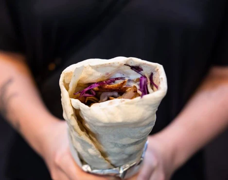 Oruc Kebap Eine Person hält eine Dönerrolle, gefüllt mit roten Kohlstreifen und weiteren Zutaten, umwickelt in Alufolie. A person is holding a kebab roll filled with red cabbage strips and other ingredients, wrapped in aluminium foil.