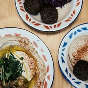 Mashery Hummus Kitchen Auf einem Tisch liegen Teller mit klassischen Mezze. Ein Teller zeigt cremigen Hummus mit Kichererbsen und frischen Kräutern, die in der Mitte angerichtet sind. Die anderen Teller enthalten Falafel-Kugeln, perfekt golden gebraten. Plates of classic mezze are laid out on a table. One plate features creamy hummus with chickpeas and fresh herbs arranged in the centre. The other plates contain falafel balls, fried to golden perfection.