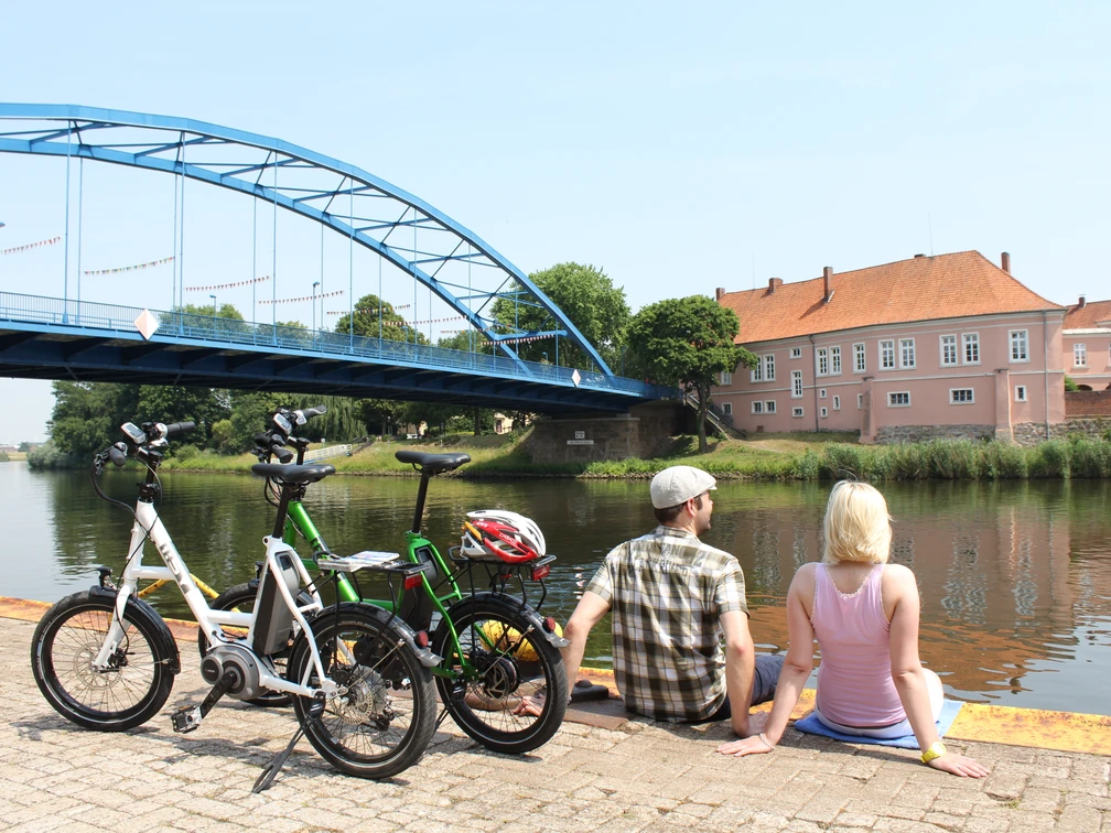Mittelweser-Region-Radfahren Zwei Personen pausieren an einem Flussufer, neben ihnen stehen Fahrräder, im Hintergrund eine Brücke.