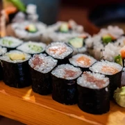 Momotaro Sushigerichte auf einem Holzbrett, inklusive Maki mit Lachs, Avocado und Gurke, kunstvoll arrangiert.Sushi dishes on a wooden board, including maki with salmon, avocado and cucumber, artfully arranged.
