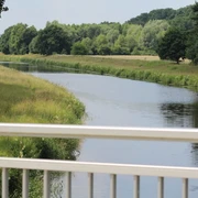 Große Aue Grüne Landschaft mit ruhigem Fluss, gesäumt von Bäumen, gesehen von einer weißen Brücke aus.