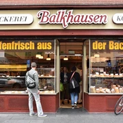Bakery Balkhausen Außenansicht auf die Bäckerei. Ein Mann schaut auf das mit Backwaren bestückte Schaufenster. Exterior view of the bakery. A man looks at the shop window filled with baked goods.