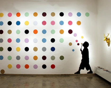30works Galerie In einem weißen Raum ist eine Installation zu sehen, die aus einer Wand mit bunten Punkten besteht. Daneben die Silhouette eines Kindes, das Blasen pustet, und eine goldene Hirschkopfskulptur.In a white room, there is an installation featuring a wall covered in colorful dots. Next to it is the silhouette of a child blowing bubbles, and a golden deer head sculpture.