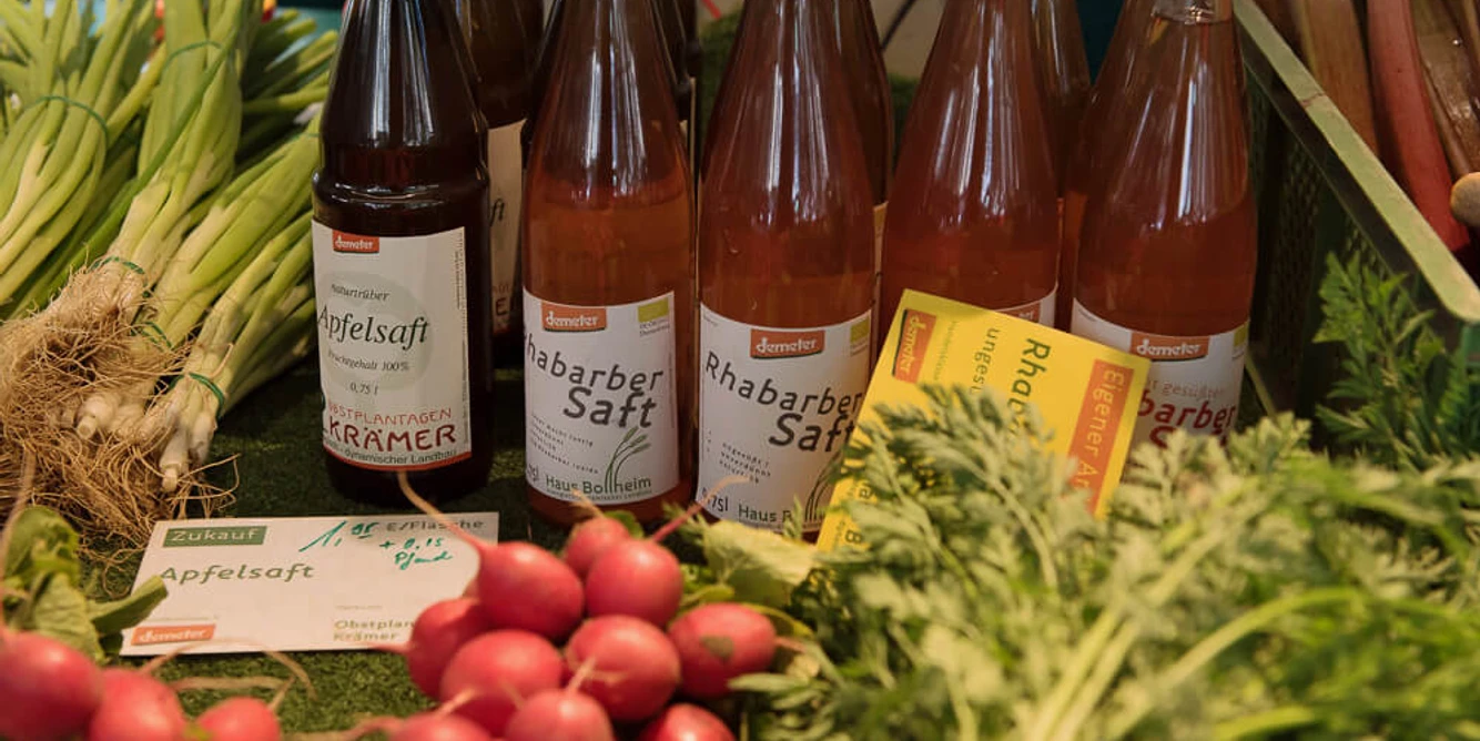 Auf einem Marktstand stehen verschiedene Flaschen Rhabarber- und Apfelsaft. Daneben frisches Gemüse wie Lauchzwiebeln, Radieschen und Rote Beete.There are various bottles of rhubarb and apple juice on a market stall. Next to them are fresh vegetables such as spring onions, radishes and beet.
