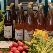Auf einem Marktstand stehen verschiedene Flaschen Rhabarber- und Apfelsaft. Daneben frisches Gemüse wie Lauchzwiebeln, Radieschen und Rote Beete.There are various bottles of rhubarb and apple juice on a market stall. Next to them are fresh vegetables such as spring onions, radishes and beet.