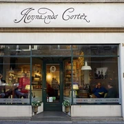 Hernando Cortez Chocolates Die Fassade der „Hernando Cortez Schokoladen“ zeigt große Schaufenster, die eine einladende Atmosphäre schaffen. Drinnen genießen Menschen den Genuss von edler Schokolade, während das stilvolle Schriftlogo über dem Eingang prangt. The façade of "Hernando Cortez Schokoladen" features large shop windows that create an inviting atmosphere. Inside, people enjoy the pleasure of fine chocolate while the stylish lettering logo is emblazoned above the entrance.