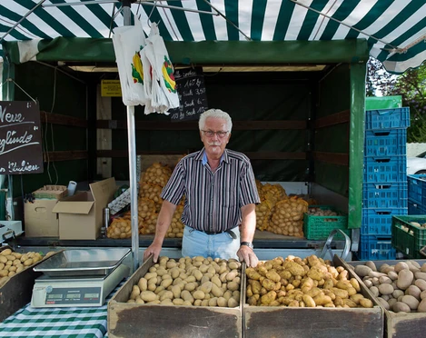 Älterer Herr verkauft frische Kartoffeln an einem Marktstand unter einem gestreiften Zelt.Elderly man selling fresh potatoes at a market stall under a striped tent.