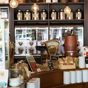 SCHAMONG KAFFEE Innenansicht eines stilvollen Cafés mit Regalen voller Kaffee- und Teedosen. Im Vordergrund sind ein Tresen mit Kaffeemaschine und viele Kaffeeartikel zu sehen.Interior view of a stylish café featuring shelves filled with coffee and tea tins. In the foreground, there's a counter with a coffee machine and various coffee-related items.