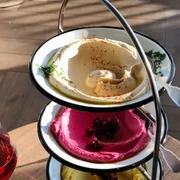 Restaurant Neni Ein dreistufiger Servierteller mit verschiedenen Mezze-Spezialitäten: cremiger Hummus, leuchtend pinke Rote-Bete-Creme und ein weiterer Aufstrich, verziert mit Kräutern und Gewürzen. Daneben ein Glas Rotwein auf einem hölzernen Tisch.A three-tiered serving platter with various mezze specialities: creamy hummus, bright pink beetroot cream and another spread, garnished with herbs and spices. Next to it, a glass of red wine on a wooden table.