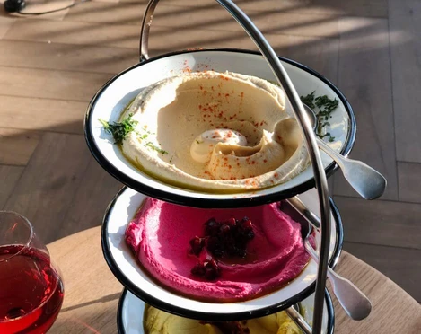 Restaurant Neni Ein dreistufiger Servierteller mit verschiedenen Mezze-Spezialitäten: cremiger Hummus, leuchtend pinke Rote-Bete-Creme und ein weiterer Aufstrich, verziert mit Kräutern und Gewürzen. Daneben ein Glas Rotwein auf einem hölzernen Tisch.A three-tiered serving platter with various mezze specialities: creamy hummus, bright pink beetroot cream and another spread, garnished with herbs and spices. Next to it, a glass of red wine on a wooden table.