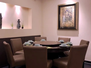 Tang Wang Ein großer runder Tisch, umgeben von gemütlichen, hellbraunen Sesseln, wird von sanftem Licht beleuchtet. Schlichte Vasen und Pflanzen in einer Nische fügen ein dezentes, dekoratives Element hinzu.A large round table surrounded by cozy, light brown armchairs, gently illuminated with soft lighting. Simple vases and plants placed in a niche add a subtle decorative touch.