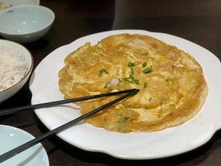 Tang Wang Goldbraunes Omelett mit Frühlingszwiebeln auf einem weißen Teller, daneben Essstäbchen.Golden-brown omelette garnished with spring onions served on a white plate, accompanied by chopsticks.