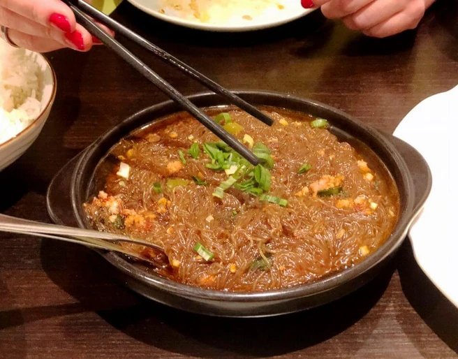 Tang Wang Ein schwarzer Teller beherbergt ein köstliches Gericht aus durchscheinenden Glasnudeln, garniert mit frischen Frühlingszwiebeln. Eine Hand hält Essstäbchen. Im Hintergrund sind Reis und andere Teller zu sehen.A black plate presenting a delicious dish of translucent glass noodles, topped with fresh spring onions. A hand is holding chopsticks. Rice and other plates are visible in the background.