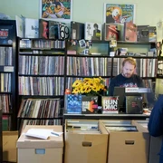 Underdog Recordstore In einem kleinen Plattenladen stehen eng gestellte Regale voller Schallplatten. Ein Verkäufer, umgeben von bunten Plattencovern und Postern an den Wänden, steht hinter einer Theke mit Kasse. Vor der Theke befinden sich Kartons mit weiteren Schallplatten.In a small record store, shelves are crammed full of records. A sales assistant, surrounded by colorful record covers and posters on the walls, stands behind a counter with a cash register. In front of the counter are boxes with more records.