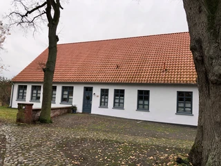Küsterhaus Daverden Ein traditionelles Gebäude mit rotem Ziegeldach und mehreren blauen Fenstern, umgeben von Bäumen.