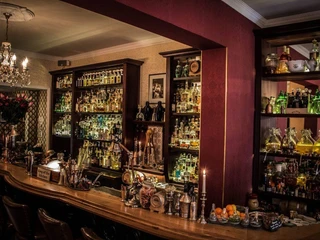 Seiberts Cocktailbar Eine elegante Cocktailbar mit langer Theke, beleuchtet von warmen Lampen, die verschiedene Spirituosen in Regalen präsentieren. Die Kerzen auf der Theke schaffen eine gemütliche Stimmung, mit Obst und Barkeeper-Tools als Dekoration.An elegant cocktail bar featuring a long counter illuminated by warm lights, showcasing various spirits arranged on shelves. Candles on the counter create a cozy ambiance, complemented by decorative fruit and bartending tools.