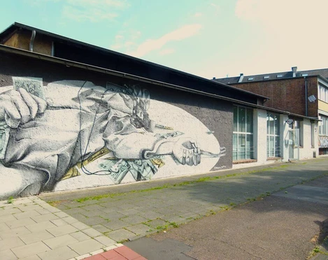 artrmx e. V. | Atelierzentrum Ehrenfeld Auf einer grauen Wand befindet sich ein beeindruckendes Wandbild. Es zeigt eine detailreiche Zeichnung eines aus Papier bestehenden Schiffes, das durch eingebettete Geldnoten segelt. Die Umgebung ist urban und ruhig, mit angrenzenden Gebäuden im Hintergrund.There is an impressive mural on a gray wall. It shows a detailed drawing of a ship made of paper sailing through embedded banknotes. The surroundings are urban and calm, with adjacent buildings in the background.