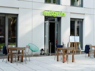 Sattgrün Außenansicht eines modernen veganen Restaurants mit heller Fassade, stilvollen Holztischen und Stühlen auf einer großzügigen Terrasse.Exterior view of a modern vegan restaurant featuring a bright facade and stylish wooden tables and chairs arranged on a spacious terrace.