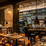 Phaedra Ein stilvoll eingerichtetes Restaurant mit gemütlicher Atmosphäre, freiliegenden Backsteinwänden und sorgfältig arrangierten Weinen in Regalen. Helle Beleuchtung akzentuiert den Barbereich, während Holztische und Stühle zum Verweilen einladen.A stylishly furnished restaurant with a cosy atmosphere, exposed brick walls, and thoughtfully arranged wines displayed on shelves. Bright lighting accentuates the bar area, while wooden tables and chairs invite guests to linger.