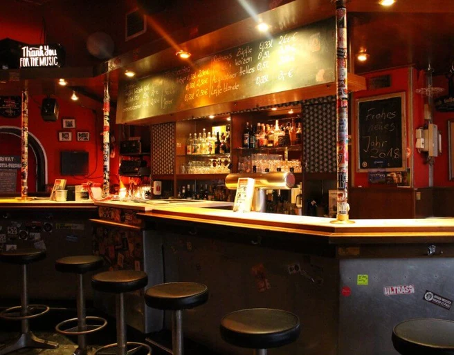 Lotta Bar Innenansicht auf den Barbereich. Die Theke ist schwarz lackiert und davor stehen Metallhocker. Interior view of the bar area. The counter is painted black and metal stools are placed in front of it.