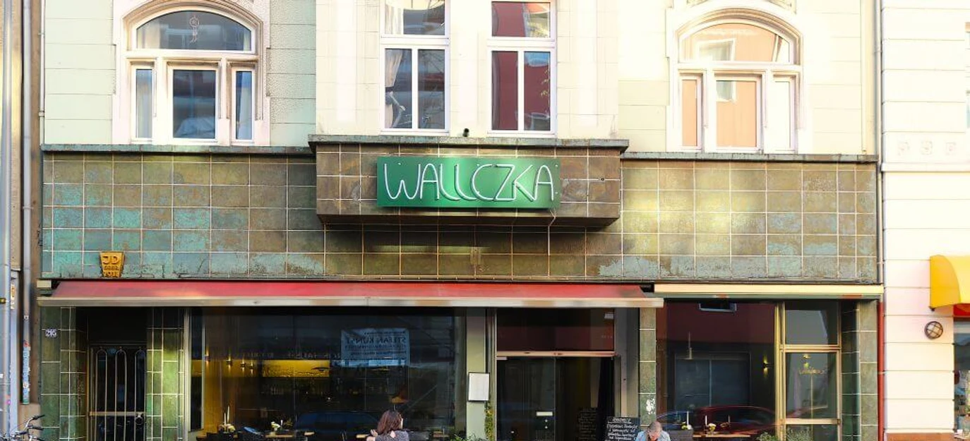 Wallczka Die Straßenszene zeigt das Restaurant Wallczka mit großen Fenstern, zwei Gästen und einem grünen Neonschild.The street scene depicts the restaurant Wallczka with large windows, two guests, and a green neon sign.