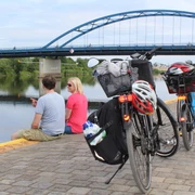 Radler Zwei Fahrräder stehen am Ufer, während ein Paar den Ausblick auf eine Brücke über einem Fluss genießt.