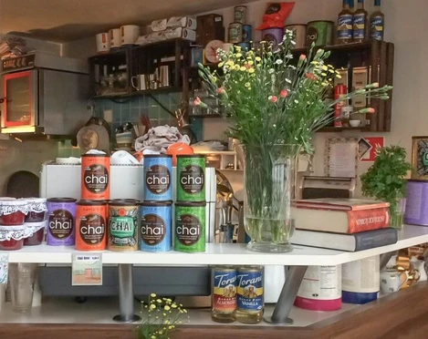 Leuchte coffee bar Cafétheke bestückt mit Marmeladengläsern, Blumen in einer Kristallvase, Chai-Tee Dosen und Büchern. Im Hintergrund hängen Weinkästen gefüllt mit Tassen und Tellern.Coffee bar stocked with jam jars, flowers in a crystal vase, chai tea tins, and books. Wine racks filled with cups and plates hang in the background.