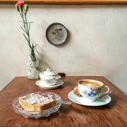 Leuchte coffee bar Auf einem Holztisch stehen eine Tasse Kaffee und ein Teller mit Mandelkuchen. Das Geschirr ist aus Porzellan und floral verziert.There is a cup of coffee and a plate of almond cake on a wooden table. The tableware is porcelain and decorated with flowers.