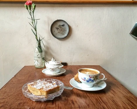 Leuchte Kaffeebar Auf einem Holztisch stehen eine Tasse Kaffee und ein Teller mit Mandelkuchen. Das Geschirr ist aus Porzellan und floral verziert.There is a cup of coffee and a plate of almond cake on a wooden table. The tableware is porcelain and decorated with flowers.