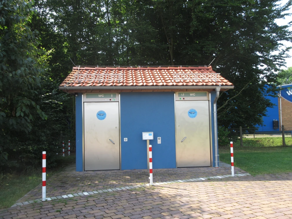 Wassarium Drakenburg Zwei moderne, barrierefreie öffentliche Toiletten mit blauen Türen in einem parkähnlichen Umfeld.
