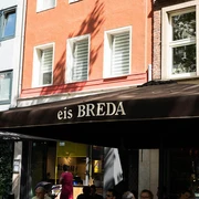 Breda Ice Cream Café Unter einem großen Sonnenschirm mit der Aufschrift eis Breda sitzen Menschen und essen Eis. People are sitting under a large sunshade with the inscription eis Breda and eating ice cream.