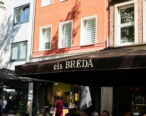 Breda Ice Cream Café Unter einem großen Sonnenschirm mit der Aufschrift eis Breda sitzen Menschen und essen Eis. People are sitting under a large sunshade with the inscription eis Breda and eating ice cream.