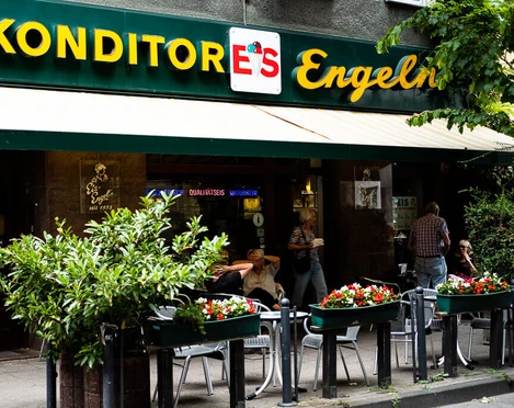 Konditorei Eis Engel Fassade mit Markise und in großen Buchstaben platziertes Schild über der Markise. Mensch sitzen vor der Konditorei.Facade with awning and sign placed in large letters above the awning. People sitting in front of the cafe.
