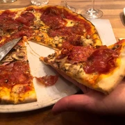 Toscanini Eine Hand hält ein Stück Pizza mit Salami und Schinken von einer angerichteten, großen Pizza mit knusprigem Rand ab.A hand holds a slice of pizza topped with salami and ham, lifted from a large, freshly prepared pizza with a crispy crust.