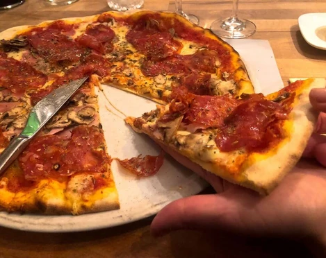 Toscanini Eine Hand hält ein Stück Pizza mit Salami und Schinken von einer angerichteten, großen Pizza mit knusprigem Rand ab.A hand holds a slice of pizza topped with salami and ham, lifted from a large, freshly prepared pizza with a crispy crust.