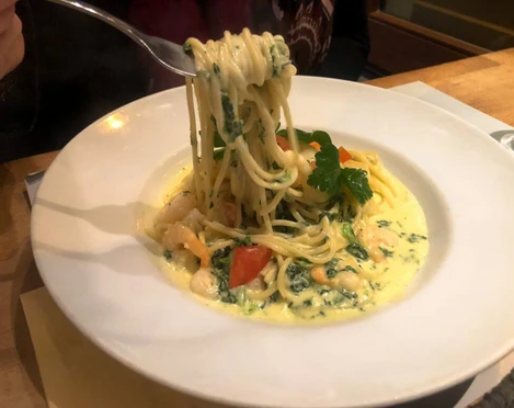 Toscanini Ein Teller Spaghetti in cremiger Soße mit Garnelen, Tomatenstücken und Petersilie auf Holzuntergrund.A plate of spaghetti in a creamy sauce with shrimp, tomato pieces, and parsley, served on a wooden surface.