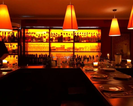 Tapeo and co Das Bild zeigt eine gemütliche Bar-Atmosphäre mit warmem orangefarbenem Licht, das von stilvollen Pendelleuchten ausgeht und die Rückwand, gefüllt mit Flaschen verschiedener Getränke, sanft erhellt. Auf den Tischen brennen ruhige Kerzen.The image depicts a cozy bar atmosphere illuminated by warm, orange-hued lighting emitted from stylish pendant lamps, softly highlighting the back wall filled with bottles of various beverages. Calmly flickering candles adorn the tables.