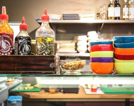 Veedel Vital Bunte Schalen stapeln sich in einem Regal einer Küche, flankiert von Flaschen mit Soßen.Colorful bowls stacked on a kitchen shelf, flanked by bottles of sauces.