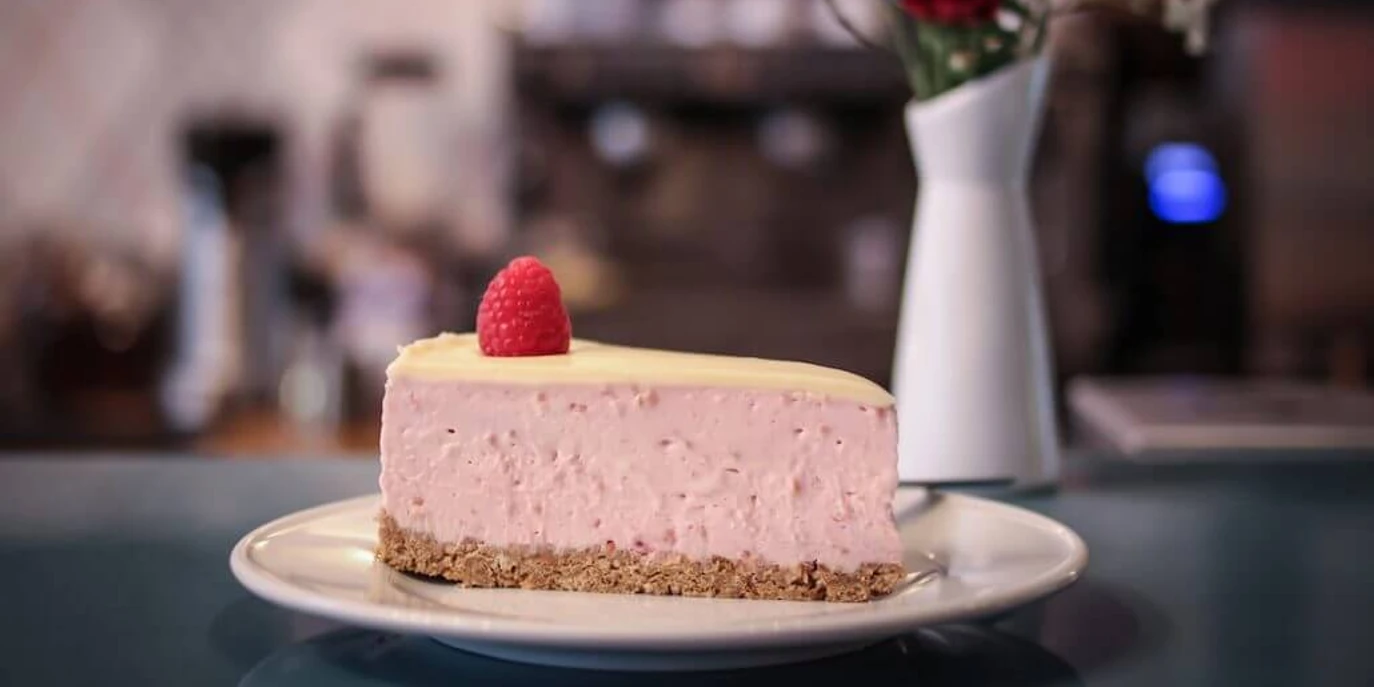 Wo ist Tom? Ein Stück Himbeer-Käsekuchen mit einer frischen Himbeere als Dekoration auf einem Teller. Im Hintergrund sind verschwommen eine Blumenvase und eine Kaffeemaschine zu erkennen.A slice of raspberry cheesecake decorated with a fresh raspberry, served on a plate. A blurred flower vase and coffee machine can be seen in the background.