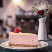 Wo ist Tom? Ein Stück Himbeer-Käsekuchen mit einer frischen Himbeere als Dekoration auf einem Teller. Im Hintergrund sind verschwommen eine Blumenvase und eine Kaffeemaschine zu erkennen.A slice of raspberry cheesecake decorated with a fresh raspberry, served on a plate. A blurred flower vase and coffee machine can be seen in the background.