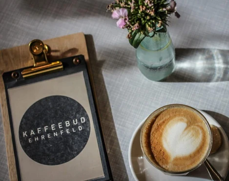 KAFFEEBUD Nahaufnahme, ein Cappuccino, daneben die Menükarte vom Café und in der Mitte eine Vase mit Blumen.Close-up of a cappuccino, the café menu next to it, and a vase of flowers in the middle.