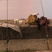 Zwei Angelruten ruhen auf einer Betonmauer am Ufer eines Sees, umgeben von Angelausrüstung.