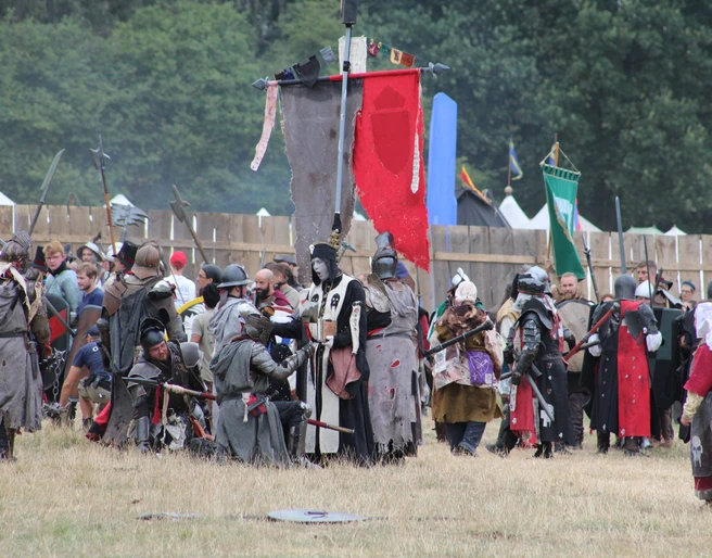 Conquest of Mythodea Menschen in mittelalterlichen Kostümen versammeln sich auf einer Wiese vor Holzzäunen und bunten Flaggen.