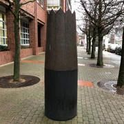 Skulptur Krone Große Skulptur einer Holz- und Metallkrone auf Pflasterweg, flankiert von Bäumen und Backsteingebäude.