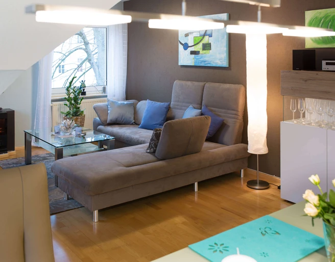 fewo-startpunkt1-wohnzimmer1.jpg Gemütliches Wohnzimmer mit grauem Sofa, Glastisch, modernen Kunstwerken und elegantem Dekor.